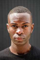 Self assured young African American male millennial in black t shirt and jeans standing on street near metal wall and looking at camera on sunny day - ADSF35455