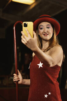 Lustiger junger weiblicher Clown mit Make-up im roten Kleid und Hut, der ein Selfie auf dem Smartphone macht, während er mit einem Stock in der Hand vor einem schwarzen Hintergrund steht - ADSF35460