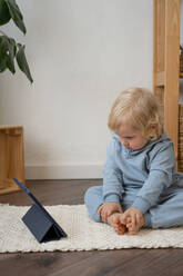 Full Body of neugierig liebenswert kleines Kind mit blonden Haaren in Freizeitkleidung sitzen auf Teppich auf dem Boden und beobachten interessante Cartoon auf Tablet in modernen Wohnung - ADSF35470