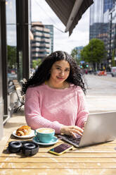 Smiling young woman with laptop sitting at sidewalk cafe on sunny day - WPEF06048