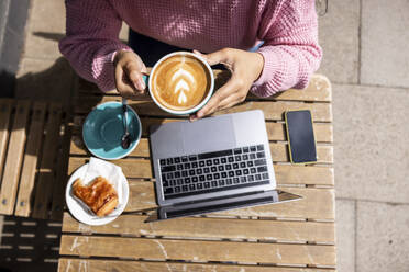 Woman holding coffee cup sitting with laptop at sidewalk cafe on sunny day - WPEF06055