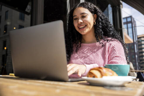Glückliche Frau mit schwarzem lockigem Haar, die einen Laptop in einem Straßencafé benutzt - WPEF06056
