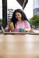 Glückliche Frau, die mit ihrem Mobiltelefon im Internet surft, sitzt mit einer Kaffeetasse in einem Straßencafé - WPEF06059