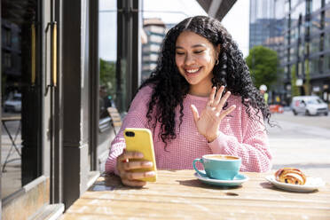 Lächelnde Frau mit winkender Hand bei einem Videoanruf über ein Smartphone in einem Straßencafé - WPEF06060