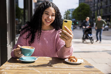 Happy woman holding coffee cup doing video call through mobile phone at sidewalk cafe - WPEF06062