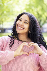 Happy young woman with black curly hair gesturing heart shape at park - WPEF06070