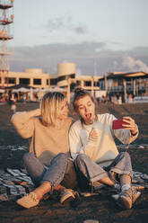 Teenager-Mädchen nimmt Selfie mit Mutter sitzt auf Picknick-Decke am Strand - OMIF00919