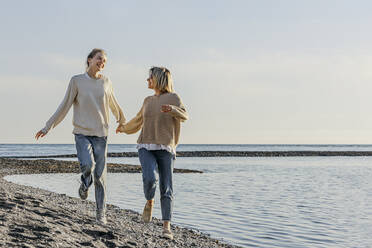 Mother and daughter holding hands running at beach - OMIF00927