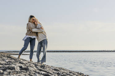 Mutter umarmt Tochter stehend am Meer am Strand - OMIF00928