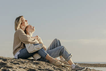 Daughter and mother sitting with eyes closed enjoying sunlight at beach - OMIF00931