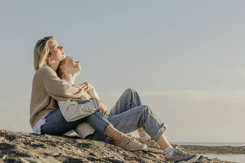 Tochter und Mutter sitzen mit geschlossenen Augen und genießen das Sonnenlicht am Strand - OMIF00931