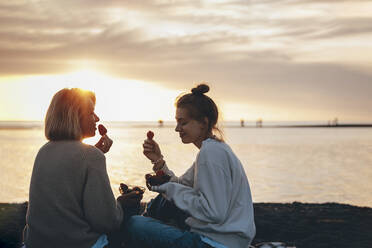 Lächelnde Mutter und Tochter essen Erdbeeren am Strand bei Sonnenuntergang - OMIF00944