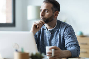 Thoughtful freelancer holding eyeglasses sitting with mug at home - JSRF02140