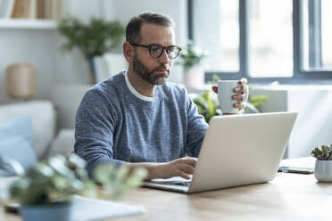 Businessman holding coffee mug working on laptop at home - JSRF02146