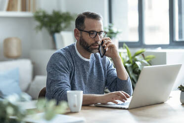 Geschäftsmann, der zu Hause am Laptop mit einem Smartphone telefoniert - JSRF02148