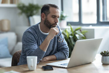 Thoughtful businessman holding eyeglasses looking at laptop working from home - JSRF02150