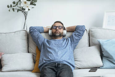 Mature man with hands behind head listening music through wireless headphones on sofa at home - JSRF02152