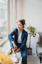 Happy businesswoman sitting by window in office - JOSEF10688