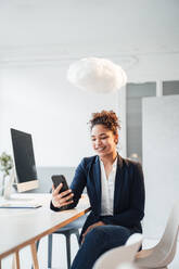 Wolke schwebt über glückliche Geschäftsfrau mit Smartphone im Büro - JOSEF10691
