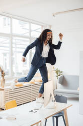 Cheerful businesswoman standing on desk in office - JOSEF10720