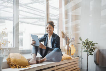 Happy young businesswoman with tablet PC sitting in front of window - JOSEF10724