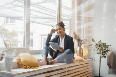 Smiling young businesswoman with tablet PC sitting in front of window - JOSEF10725