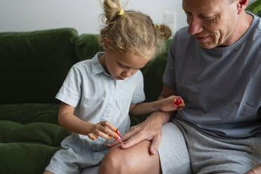 Daughter painting nails of father sitting on sofa at home - SVKF00390