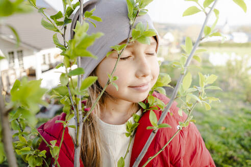 Lächelndes Mädchen mit geschlossenen Augen bei einer Pflanze im Garten - KMKF01847