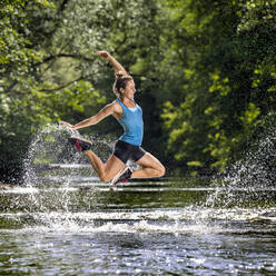 Junge Frau mit erhobener Hand, die über einen Fluss springt - STSF03257