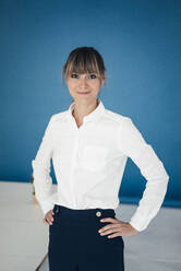 Confident businesswoman standing in front of blue wall at office - JOSEF10790