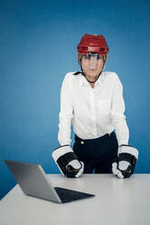 Businesswoman wearing ice hockey helmet and gloves at table in front of blue wall - JOSEF10793