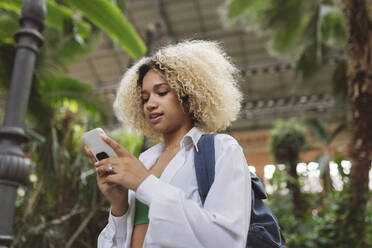 Woman with curly hair using smart phone in front of trees - JCCMF06589