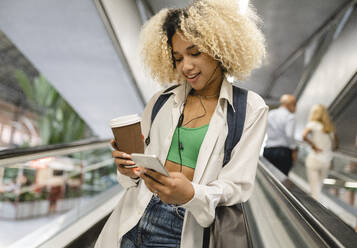 Frau mit Einwegbecher und Smartphone auf Rolltreppe am Flughafen lehnend - JCCMF06591