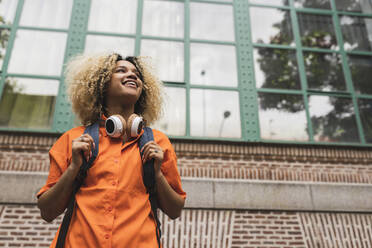 Smiling woman with headphones standing in front of building - JCCMF06613