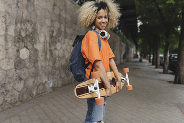 Lächelnde Frau mit Rucksack und Skateboard auf dem Fußweg - JCCMF06621