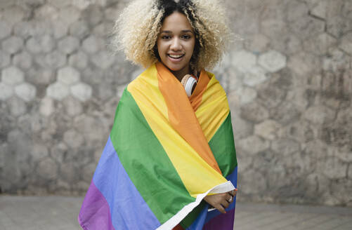 Smiling woman covered in rainbow flag standing in front of wall - JCCMF06626
