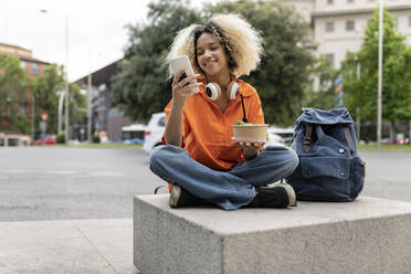 Lächelnde Frau, die ein Smartphone benutzt und eine Lunchbox hält, die im Schneidersitz auf dem Sitz steht - JCCMF06632