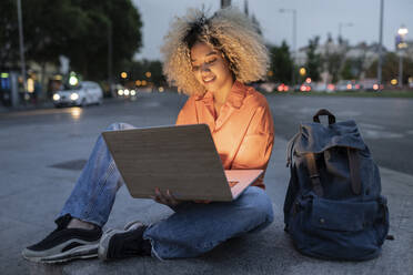 Frau mit Laptop sitzt mit Rucksack auf dem Gehweg - JCCMF06660