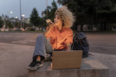 Frau mit Laptop im Gespräch über Lautsprecher auf dem Bürgersteig sitzend - JCCMF06661