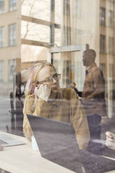 Junger nicht-binärer Unternehmer sitzt in einem Büro eines Tech-Start-ups, gesehen durch ein Glasfenster - MASF30850