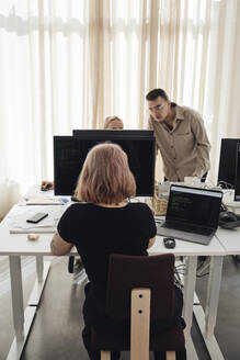 Geschäftsfrau mit Computer am Schreibtisch, während Kollegen im Büro diskutieren - MASF30856