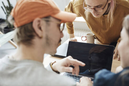 Nicht-binärer Computerprogrammierer, der auf den Laptop-Bildschirm schaut, während Kollegen bei einer Besprechung im Büro diskutieren - MASF30902