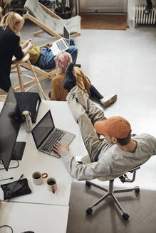 Männlicher Unternehmer mit hochgelegten Füßen auf dem Schreibtisch, der einen Laptop benutzt, während seine Kollegen im Büro eines Technologie-Start-ups diskutieren - MASF30927