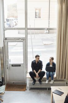 Junge Geschäftskollegen diskutieren miteinander, während sie vor einem Glasfenster im Büro sitzen - MASF30928