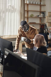 Männlicher Unternehmer, der eine Kaffeetasse hält und sich auf den Schreibtisch stützt, während er sich mit einer Kollegin im Büro unterhält - MASF30931