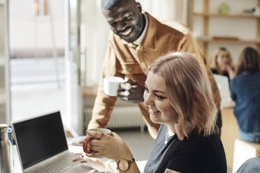 Seitenansicht einer lächelnden Unternehmerin, die mit einem Kollegen im Büro eine Kaffeetasse hält - MASF30932