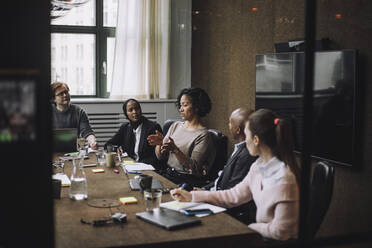 Männliche und weibliche Kollegen betrachten eine Geschäftsfrau, die am Konferenztisch sitzend Ideen austauscht, während sie gestikulieren - MASF30944