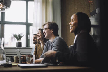 Cheerful young businessman sitting by male and female colleagues while discussing in meeting room - MASF30953