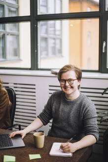 Porträt eines glücklichen jungen Geschäftsmannes, der mit Laptop und Tagebuch am Konferenztisch sitzt - MASF30975