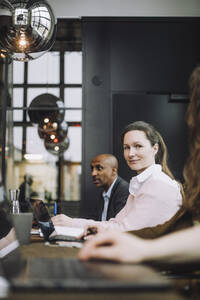 Porträt einer mittleren erwachsenen Geschäftsfrau, die mit einem Laptop neben Kollegen in einem Besprechungsraum sitzt - MASF30978